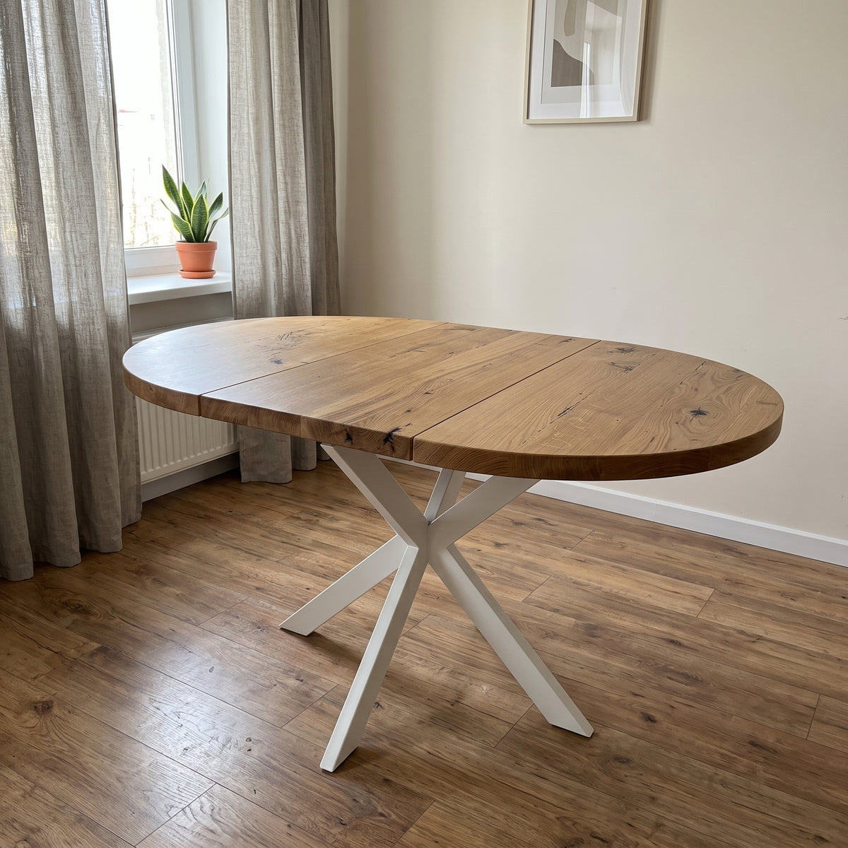 An oval wooden dining table with a natural oak top and a white painted X-shaped metal base, shown in a bright room with wood floors and a window.