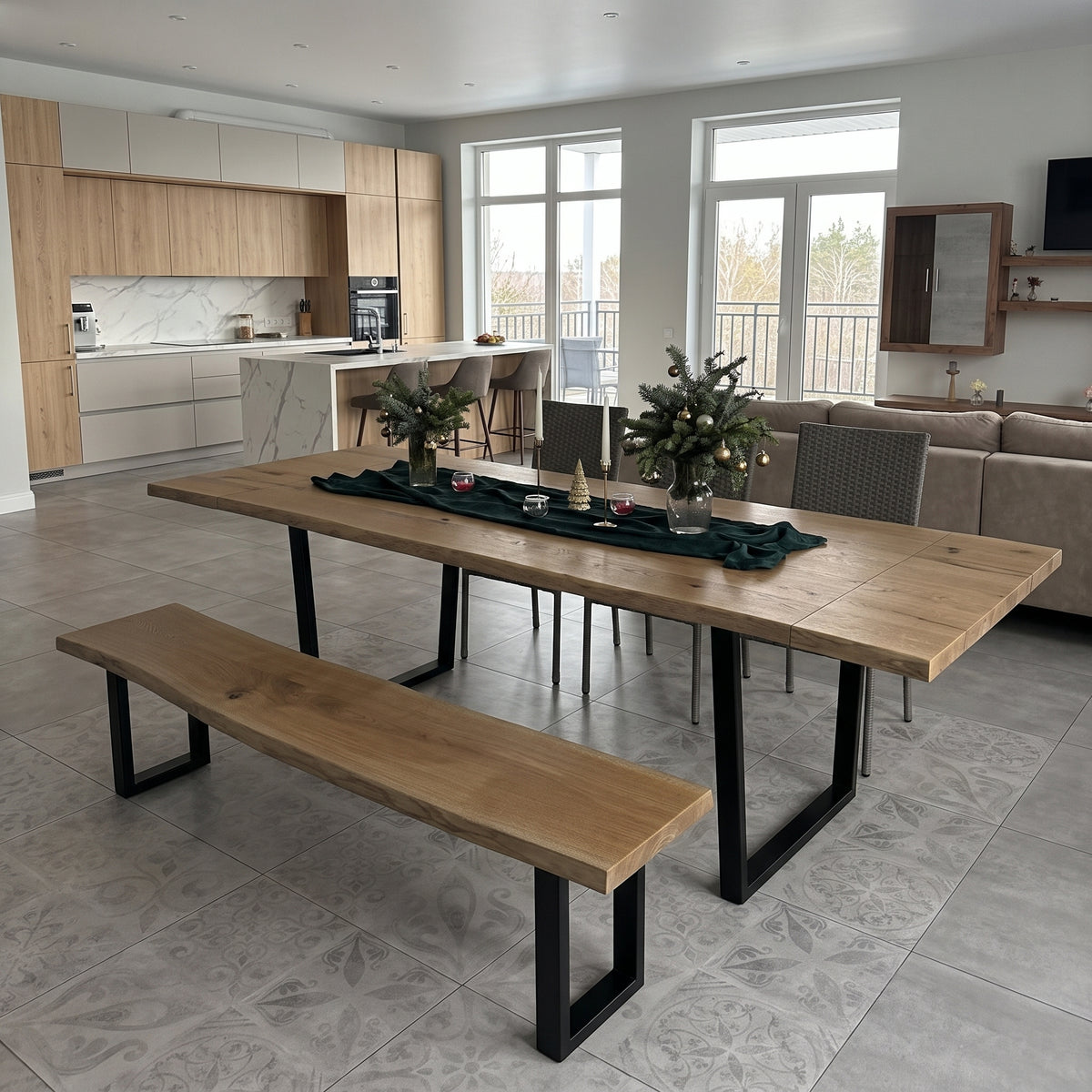 A large rectangular wooden dining table with black metal legs, accompanied by a matching wooden bench and several gray chairs, set in a modern open-plan dining room with a kitchen and living room in the background.