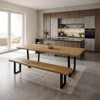 A modern dining area featuring a large rectangular wooden table with black metal legs and a matching wooden bench, set on a light patterned rug in a contemporary kitchen with concrete floors and wood cabinetry.