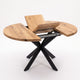 A modern round dining table with a natural wood grain top and a black metal X-shaped base, shown with the center panel raised to reveal the table's extension mechanism.