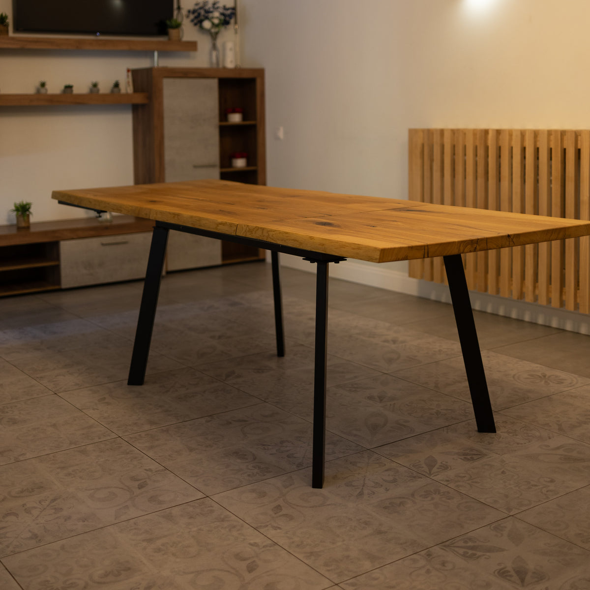 A rectangular wooden dining table with a natural oak-like top and four black metal legs, shown in a modern living room setting with tiled flooring and wooden furniture in the background.