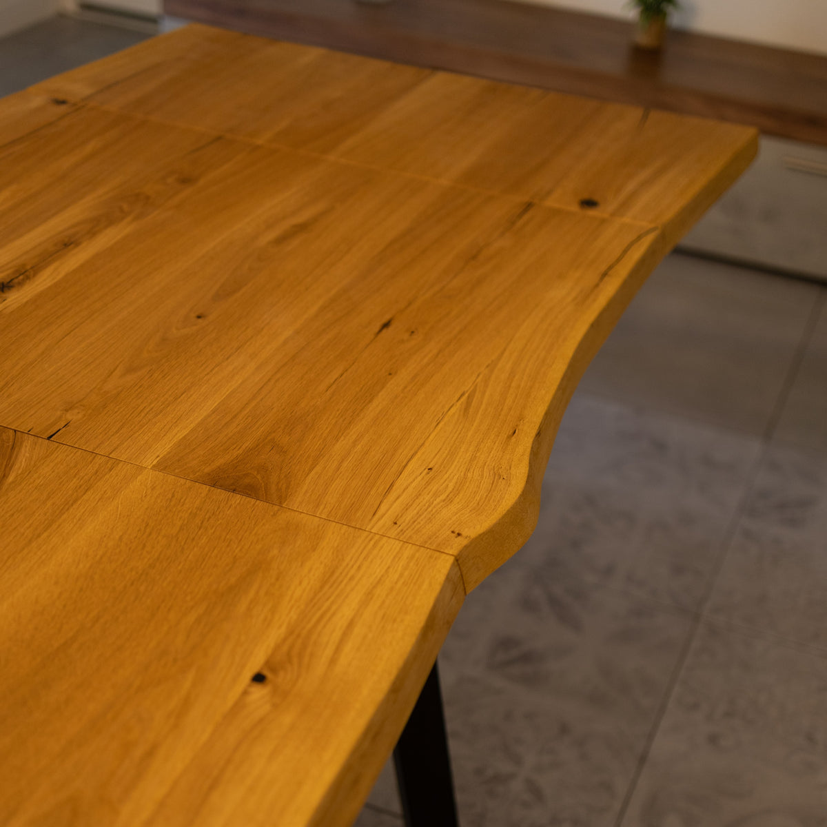 Close-up of a wooden table top with a warm honey finish and visible wood grain, shown in a real indoor setting with tiled flooring and a blurred background.