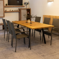 A rectangular wooden dining table with black metal legs, surrounded by six dark woven armchairs, shown in a furnished indoor room with tiled flooring and wall shelving.