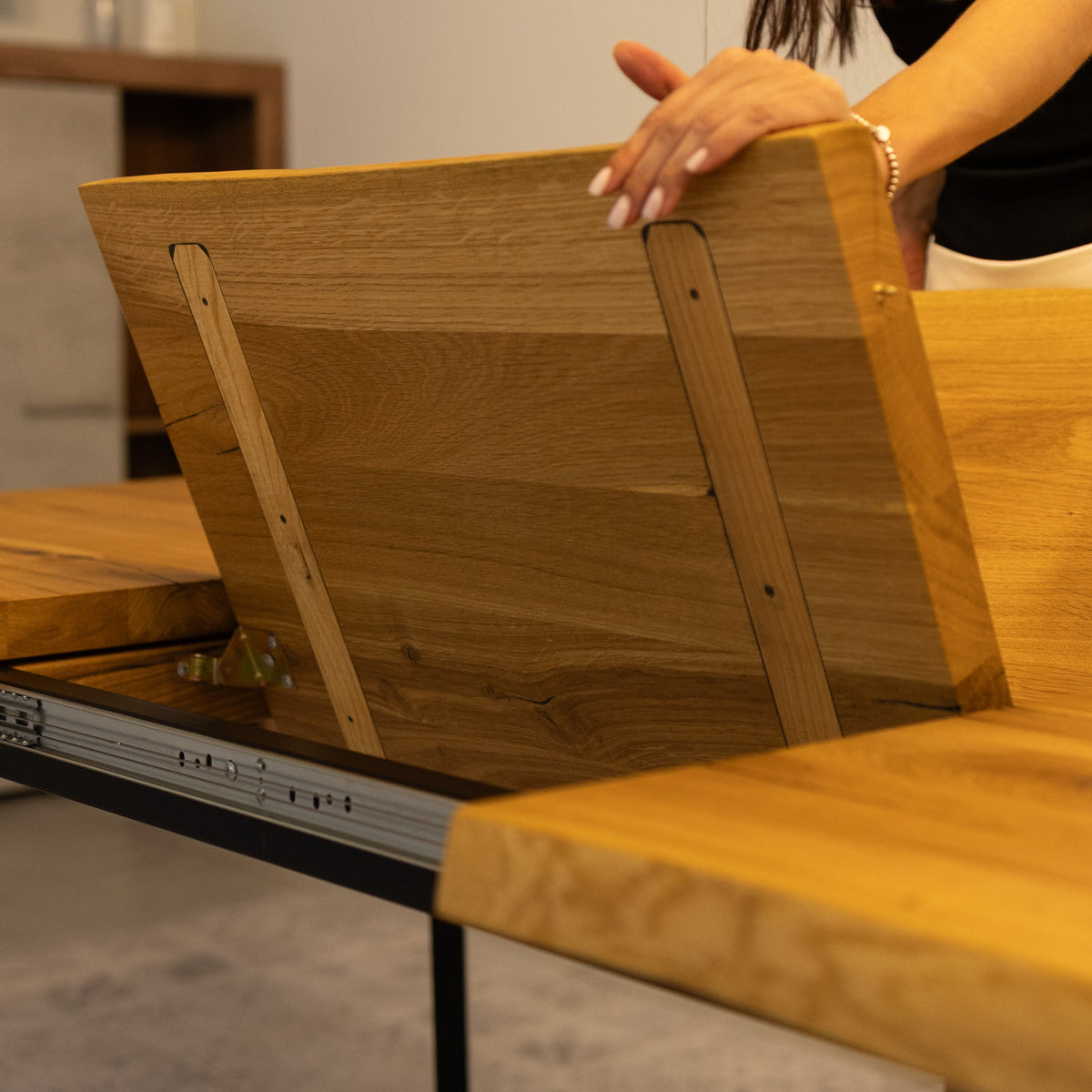 A close-up of a wooden table being opened by a person, showing the underside of the tabletop with metal support rails and a hand adjusting the panel.
