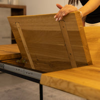 A close-up of a wooden table being opened by a person, showing the underside of the tabletop with metal support rails and a hand adjusting the panel.
