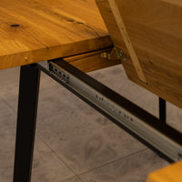 Close-up of a wooden extendable table with a black metal leg and a sliding drawer mechanism under the tabletop, set on a gray tiled floor.