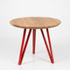 A round wooden dining table with a warm medium-brown wood grain top and three glossy red metal legs, shown on a clean white studio background.