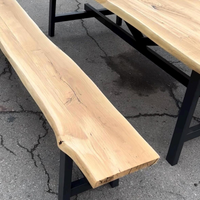 A live-edge wooden bench with a natural wood top and black metal legs, photographed outdoors on asphalt next to a matching wooden table.