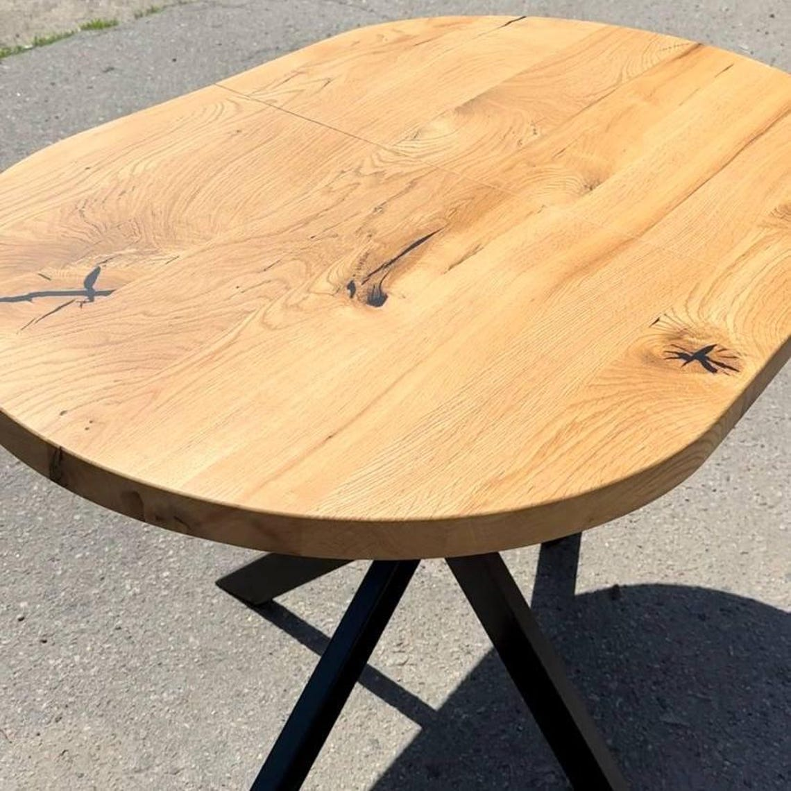A rustic oval wooden table with a natural oak top and black metal legs, photographed outdoors on a gray asphalt surface.