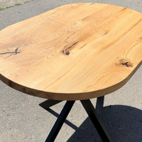 A rustic oval wooden table with a natural oak top and black metal legs, photographed outdoors on a gray asphalt surface.