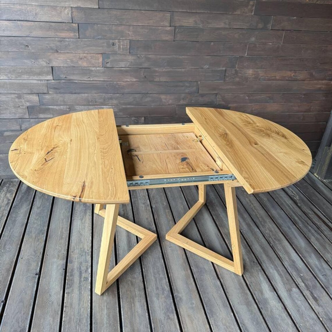 A light natural wood extendable dining table with a split oval tabletop and angled legs, shown open on a dark wood plank floor against a dark wood wall.