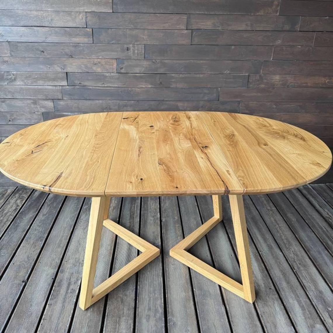 An oval wooden dining table with a natural light oak finish and geometric angled legs, set against a dark wood plank wall and floor.