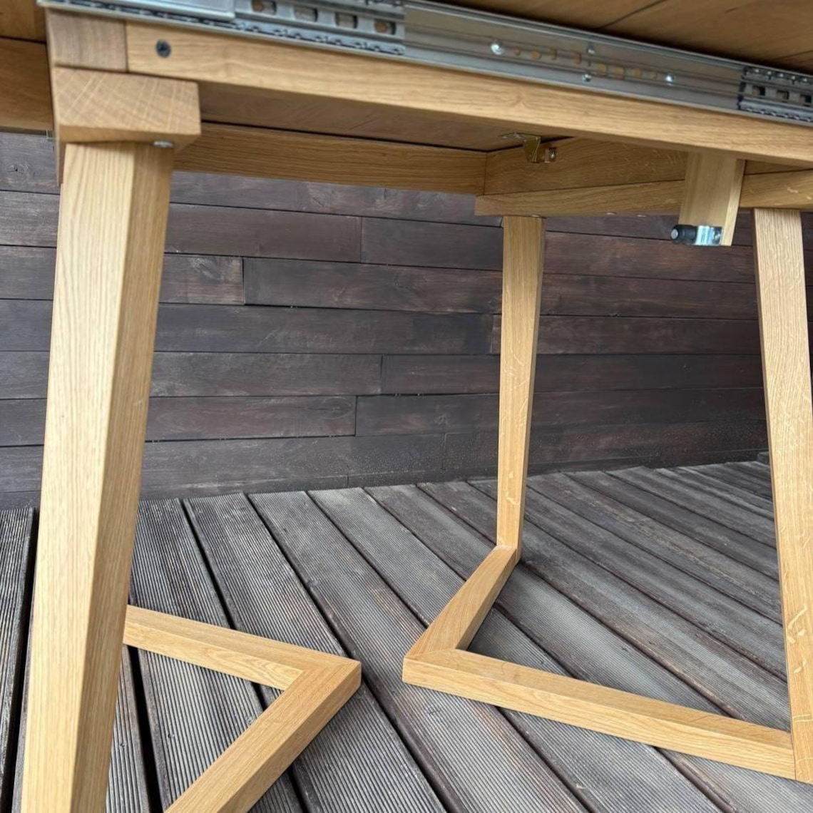 Close-up of a light natural wood table frame with metal drawer slides on a dark wood plank deck against an outdoor wall.