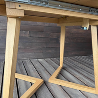 Close-up of a light natural wood table frame with metal drawer slides on a dark wood plank deck against an outdoor wall.