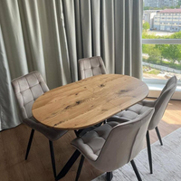 An oval wooden dining table with a natural oak finish, surrounded by four tufted gray velvet chairs with black legs, set in a bright room with a window and curtains.