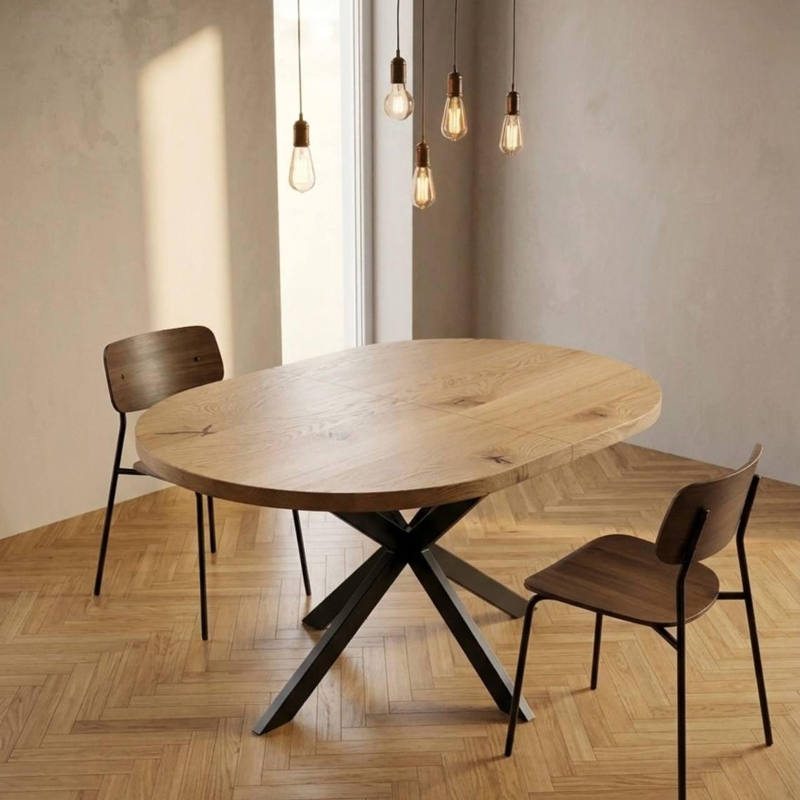 A round wooden dining table with a black metal X-shaped pedestal base, accompanied by two modern chairs with dark wood seats and black metal legs, set in a bright room with a herringbone wood floor and hanging pendant lights.