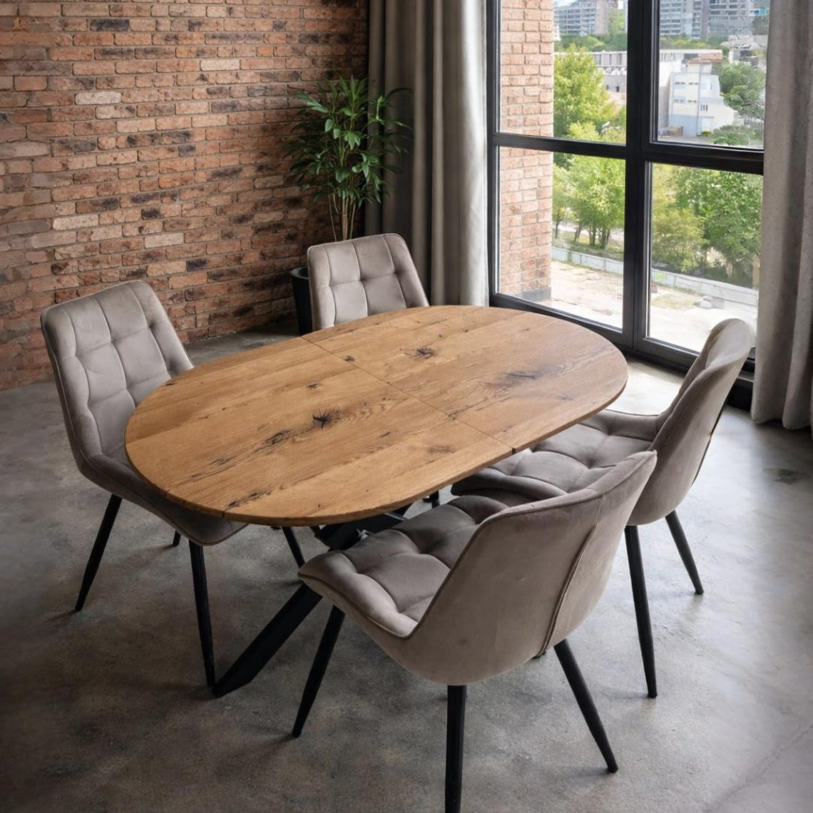 An oval wooden dining table with a natural oak finish, surrounded by four tufted upholstered chairs in a neutral gray-beige velvet fabric, set in a modern loft room with a brick wall and large windows.