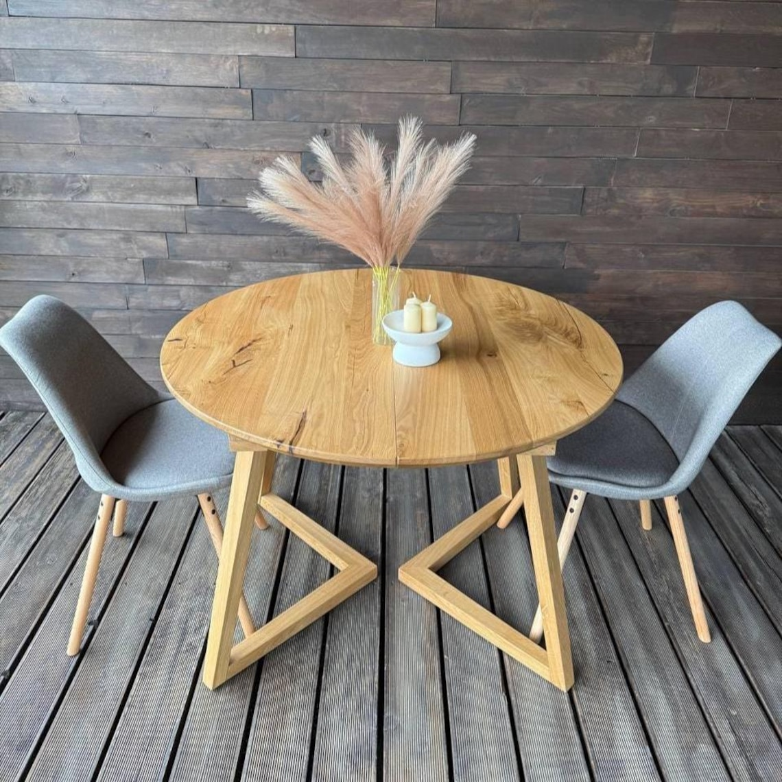 Round wooden dining table with geometric angled legs, accompanied by two gray upholstered chairs, set on a dark wood plank floor against a dark wood paneled wall. A vase with pampas grass and a candle holder sits on the table.