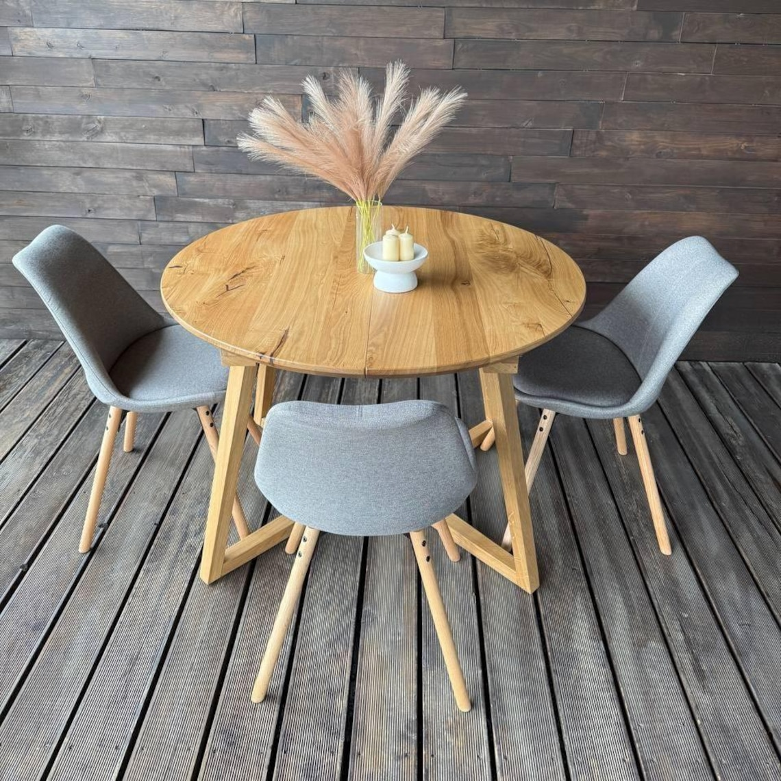 A round natural oak dining table with three gray upholstered chairs on a dark wood plank floor, set against a dark wood paneled wall. A vase with pampas grass and candles sits on the table.