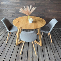 A round natural oak dining table with three gray upholstered chairs on a dark wood plank floor, set against a dark wood paneled wall. A vase with pampas grass and candles sits on the table.