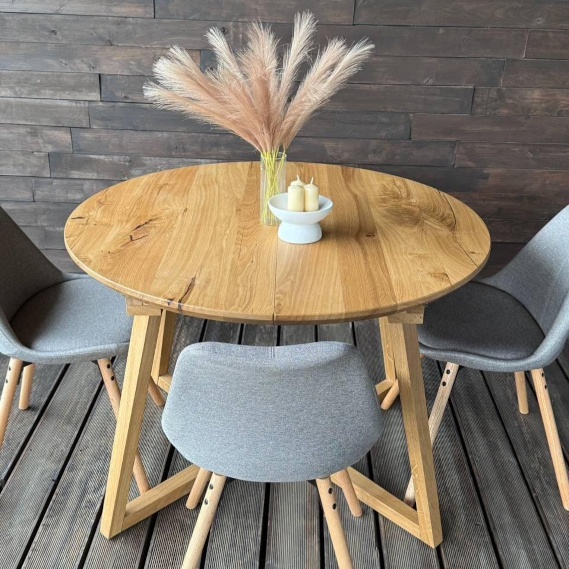 Round wooden dining table with a natural oak finish, accompanied by three gray upholstered chairs, set against a dark wood paneled wall on a wooden deck.