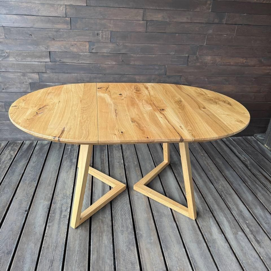 An oval wooden dining table with a natural oak finish, featuring a fold-out leaf design and geometric angled legs, set on a gray wooden deck against a dark wood paneled wall.