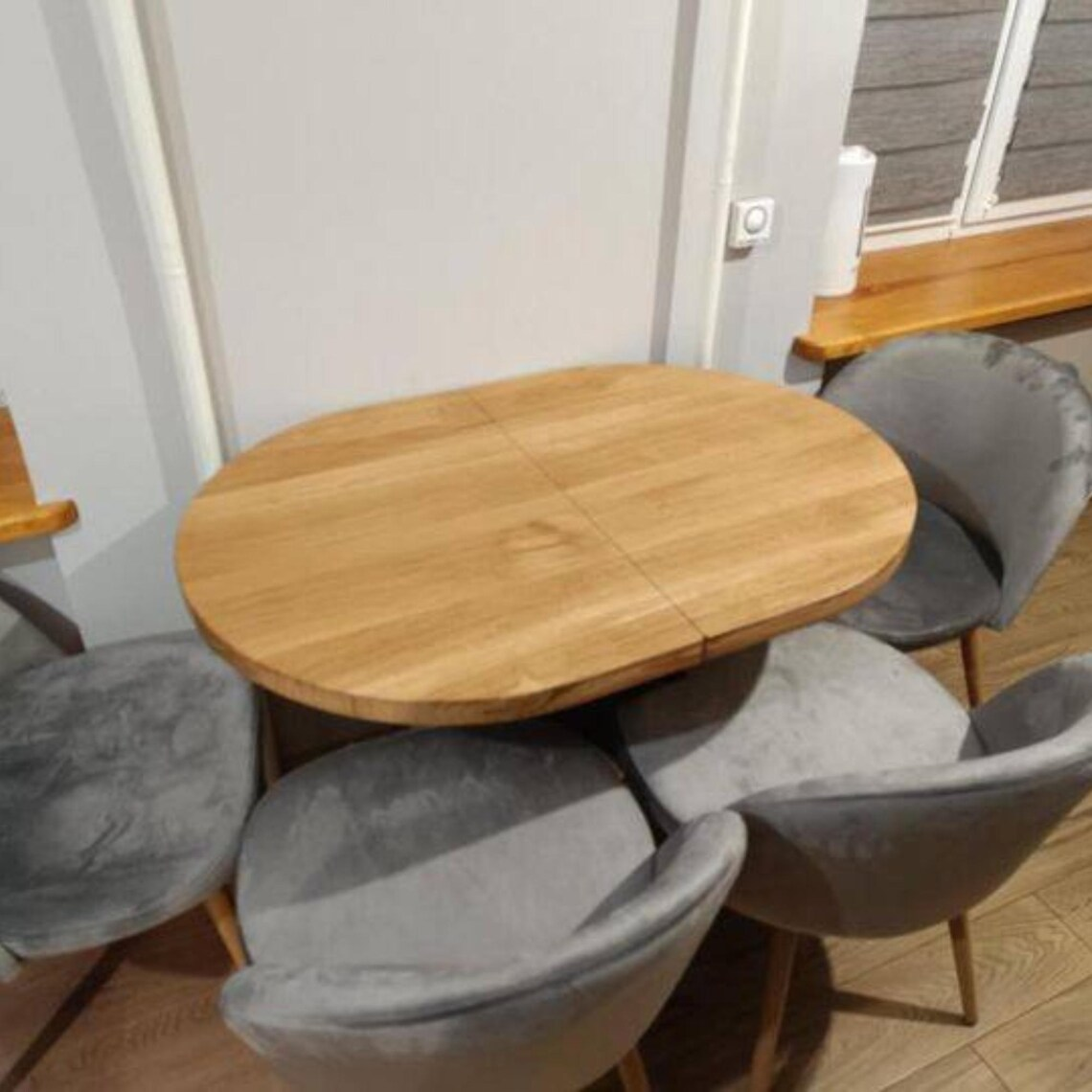 An oval wooden dining table with a light oak finish, surrounded by four gray velvet upholstered chairs with wooden legs, set in a home interior.