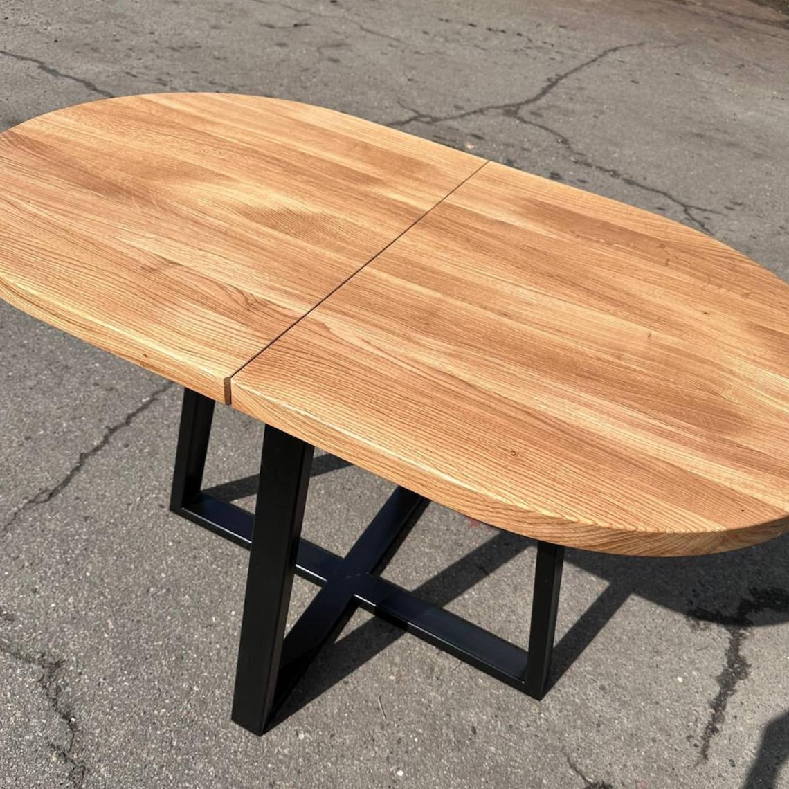 An oval wooden dining table with a natural oak finish and a black metal X-frame base, photographed outdoors on asphalt pavement.
