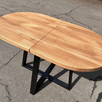 An oval wooden dining table with a natural oak finish and a black metal X-frame base, photographed outdoors on asphalt pavement.