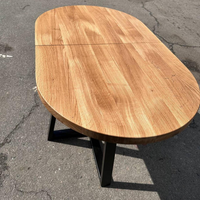 An oval wooden dining table with a light oak finish and a black metal pedestal base, photographed outdoors on asphalt pavement.