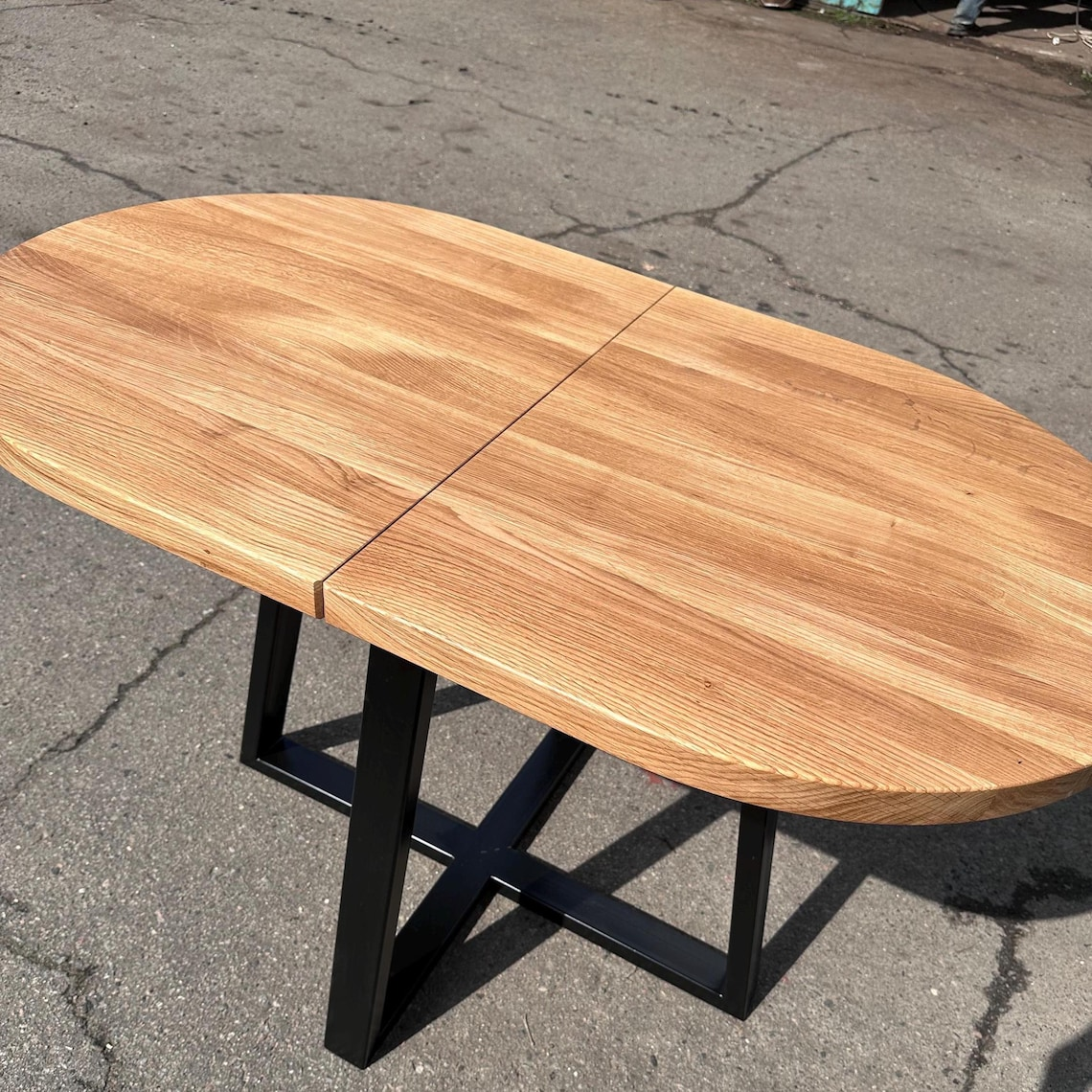 An oval wooden dining table with a natural oak finish and a black metal X-frame base, photographed outdoors on asphalt pavement.