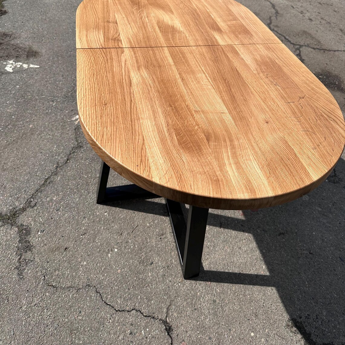 An oval wooden table with a natural oak finish and a black metal base, photographed outdoors on asphalt pavement.