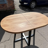 An oval wooden dining table with a light oak finish and a black metal X-frame base, photographed outdoors on asphalt with a brick wall and a blue car visible in the background.