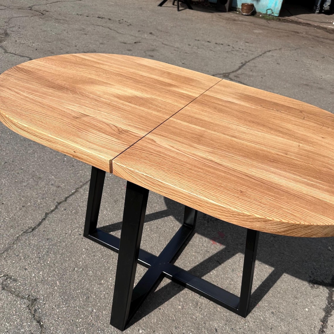 An oval wooden dining table with a natural oak finish and a black metal frame base, photographed outdoors on asphalt pavement.