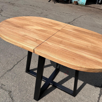 An oval wooden dining table with a natural oak finish and a black metal frame base, photographed outdoors on asphalt pavement.