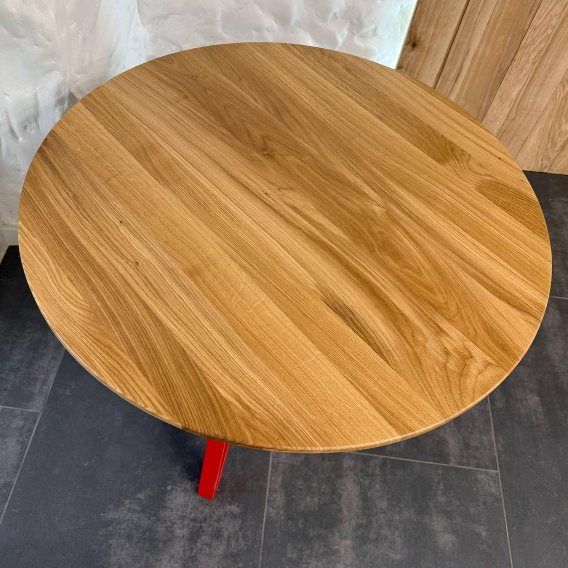 Round wooden table top with a natural oak grain finish, supported by a red metal leg, shown from an overhead angle on a dark tiled floor.