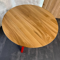 Round wooden table top with a natural oak grain finish, supported by a red metal leg, shown from an overhead angle on a dark tiled floor.