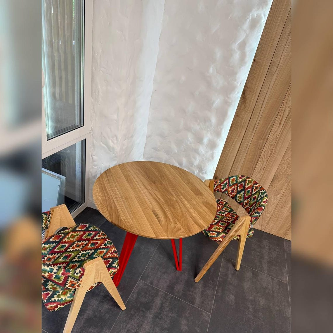 A round wooden dining table with red legs, paired with two patterned upholstered chairs, set in a modern corner room with a textured white wall, wood paneling, and dark gray flooring.