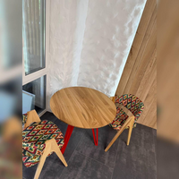 A round wooden dining table with red legs, paired with two patterned upholstered chairs, set in a modern corner room with a textured white wall, wood paneling, and dark gray flooring.