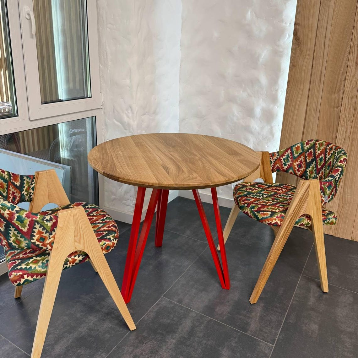 A round wooden dining table with red metal legs, paired with two wooden chairs featuring colorful geometric patterned upholstery, set in a modern room with gray tile flooring and a textured white wall.