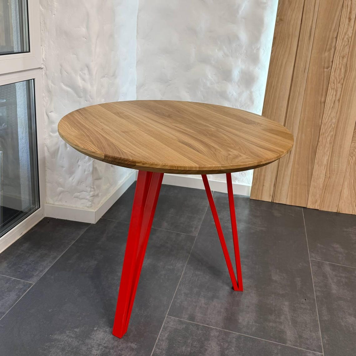 A round wooden dining table with a natural oak finish and vibrant red metal hairpin legs, set on a dark gray tiled floor against a white textured wall.