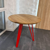 A round wooden dining table with a natural oak finish and vibrant red metal hairpin legs, set on a dark gray tiled floor against a white textured wall.