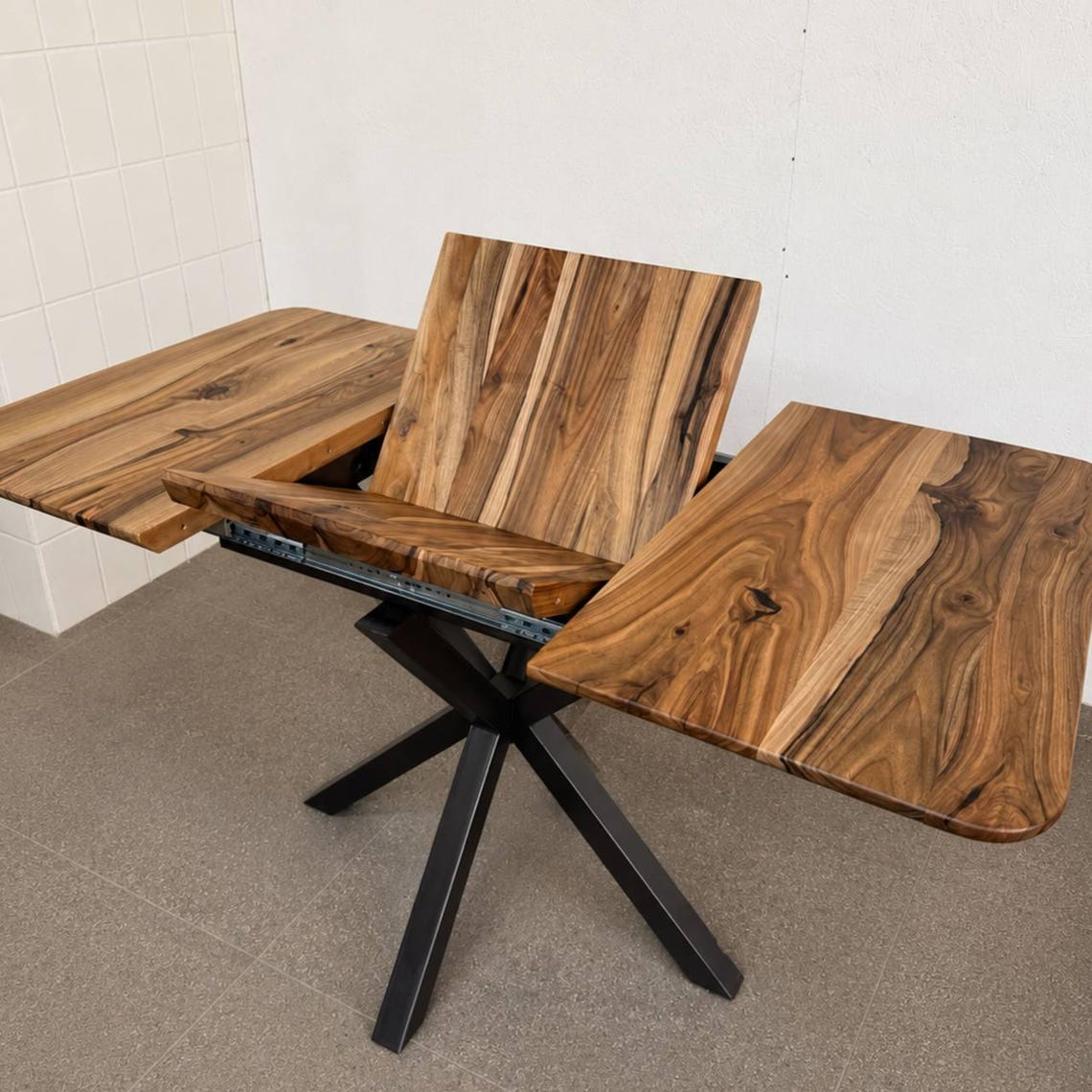 A rustic wooden dining table with a black metal X-frame base, shown with its center panel raised and side panels extended. The table features rich wood grain with prominent black knots and natural live edges, photographed in a warehouse or workshop setting with a tiled corner wall and concrete floor.