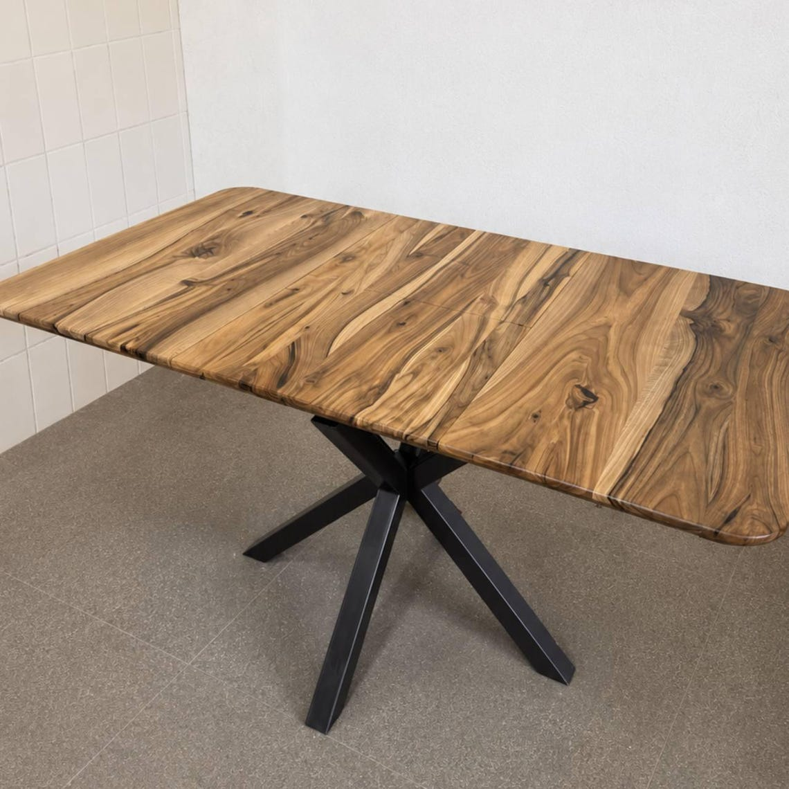 A rectangular wooden dining table with a rustic live-edge wood top and a black metal X-shaped pedestal base, shown in a room with tiled walls and a gray floor.