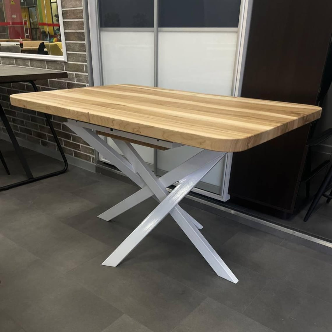 A rectangular wooden dining table with a light natural wood top and a white metal X-shaped base, shown in an indoor showroom setting.