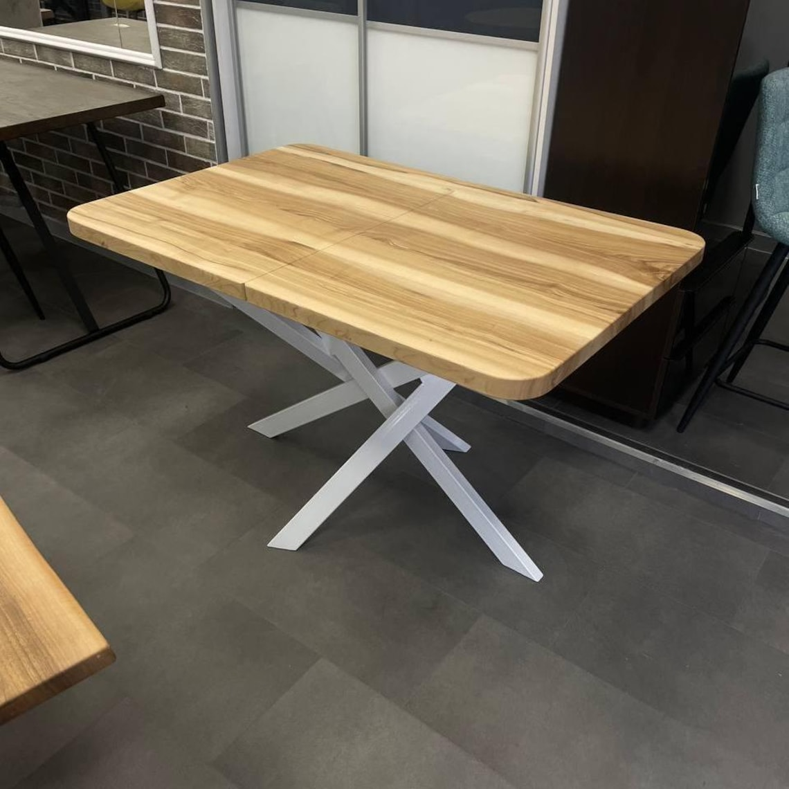 A rectangular wooden dining table with a light oak finish and a white cross-shaped metal base, shown in an indoor showroom setting.