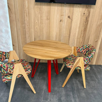 A round wooden dining table with bright red legs, surrounded by two modern chairs with colorful geometric patterned upholstery and light wood legs, set against a wood-paneled wall.