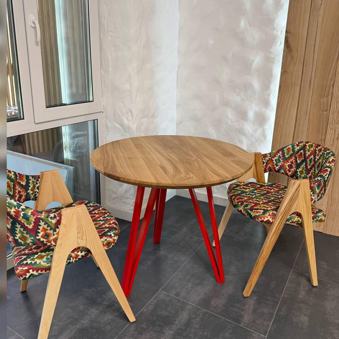 A round wooden dining table with red metal legs, accompanied by two wooden chairs with colorful geometric patterned upholstery, set in a modern room with dark gray tiled floors and textured white walls.