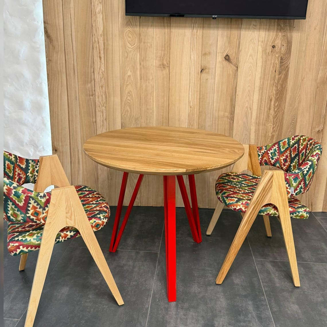 A round wooden dining table with bright red legs, surrounded by two modern wooden chairs with colorful geometric patterned upholstery, set against a wood-paneled wall.