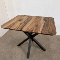 A square wooden dining table with a rustic live-edge wood top and a black metal X-shaped pedestal base, photographed in a simple indoor setting.